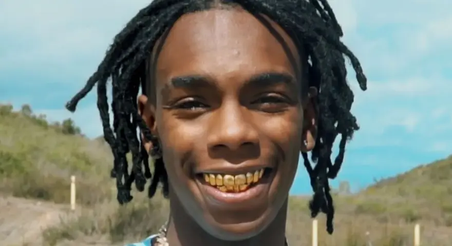 YNW Melly, uśmiechnięty z złotymi zębami, na tle nieba i wzgórz.