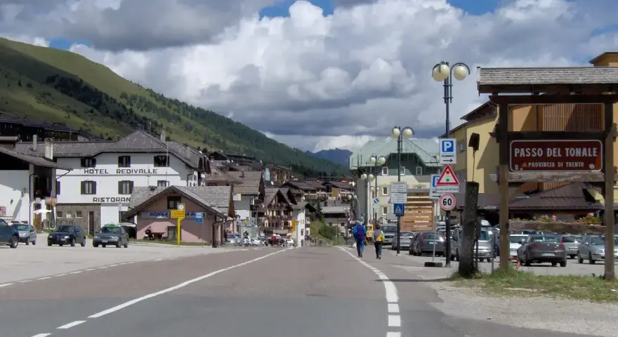 Strada verso il Passo del Tonale in moto, con case alpine e montagne sullo sfondo.