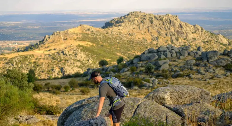 Un excursionista con mochila asciende por un terreno rocoso en la ruta Las Machotas, con un paisaje montañoso y un valle al fondo.