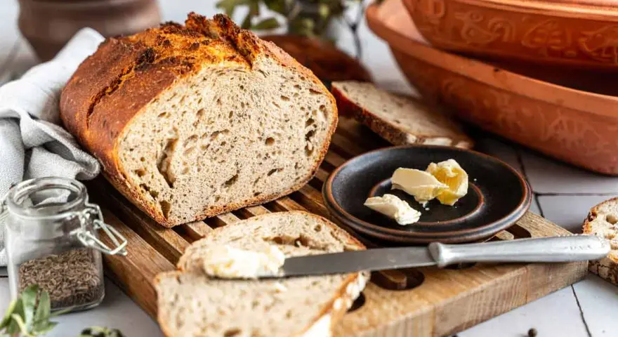 Ein rustikales Roggenbrot mit Sauerteig, aufgeschnitten und mit Butter bestrichen. Daneben ein Glas Kümmel und ein Messer.