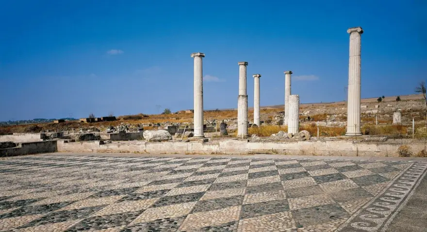 Ruines antiques avec colonnes imposantes et un sol en mosaïque de galets, évoquant l'origine d'Alexandre le Grand.