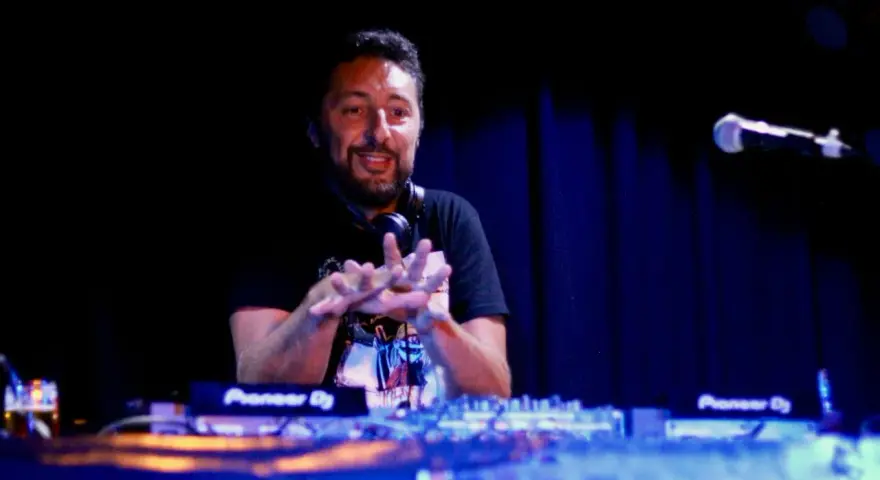 Un dj hip hop souriant, mains en l'air, mixe sur scène devant un public invisible.