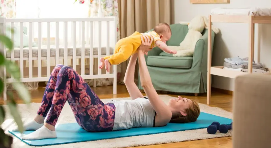 Frau macht Beckenbodentraining mit Baby. Sie liegt auf einer Matte und hebt ihr Kind hoch.