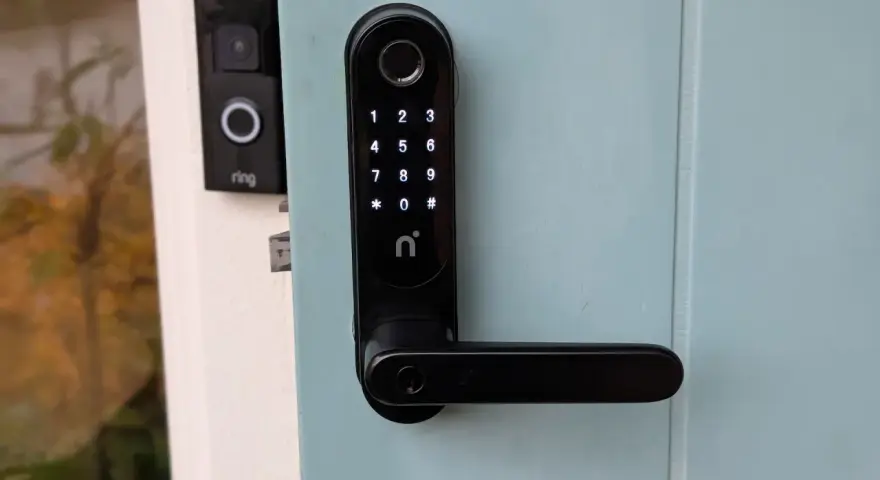 Nimly Touch Pro test: Svart digital dörrlås med kodpanel och fingeravtrycksläsare monterat på en ljusblå dörr. Bredvid syns en Ring-ringklocka.