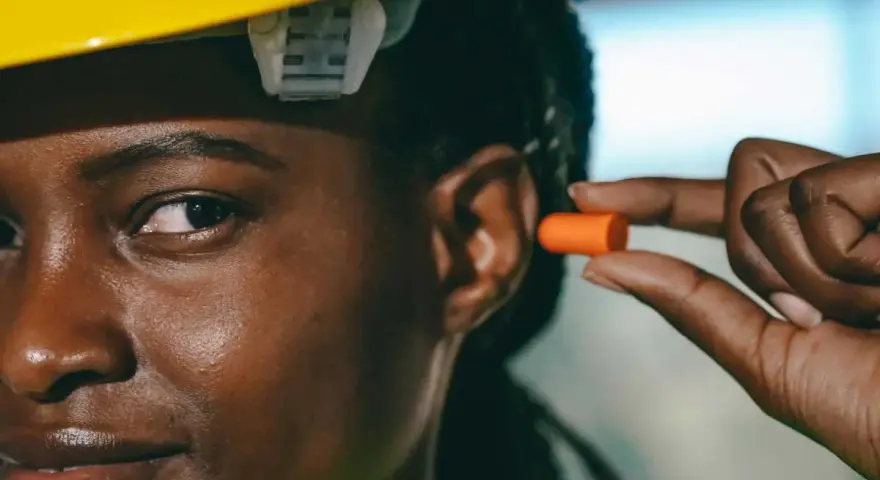 Une femme porte un casque jaune et s'apprête à insérer un bouchon d'oreille orange pour éviter le sifflement.