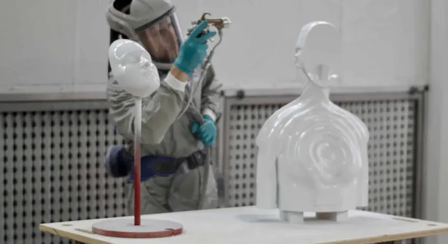 A person in a protective suit sprays a white 3D printed bust and torso, demonstrating how to paint 3d prints for a smooth finish.