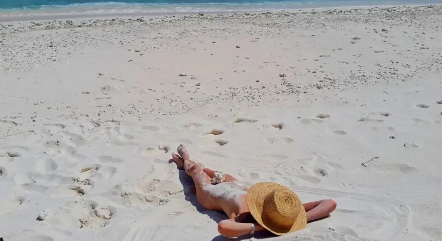 Słoneczna plaża, błękitne morze i osoba opalająca się na piasku. W tle widać fale i biały piasek z licznymi śladami stóp.