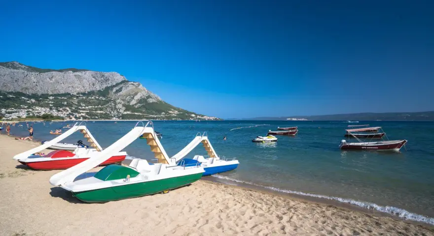 Plaża z łódkami i zjeżdżalniami, idealna na wakacje w camping Galeb. W tle góry i turkusowe morze.