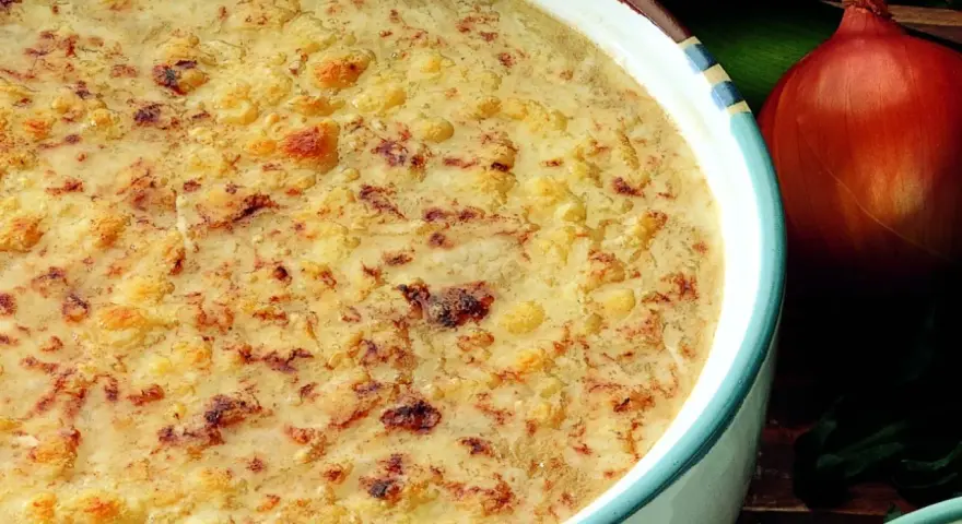 Una ricca zuppa di cipolle gratinata, con crostini dorati e formaggio fuso. Accanto, una cipolla rossa e foglie verdi.