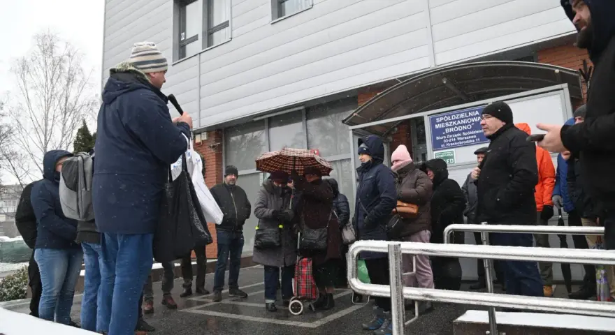 Grupa ludzi stoi przed budynkiem spółdzielni mieszkaniowej w deszczu, niektórzy pod parasolem.