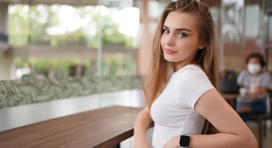 Uśmiechnięta blondynka w białej bluzce, z zegarkiem na ręku, siedzi przy stoliku. Czy to jej sekret, jak uwieść faceta?
