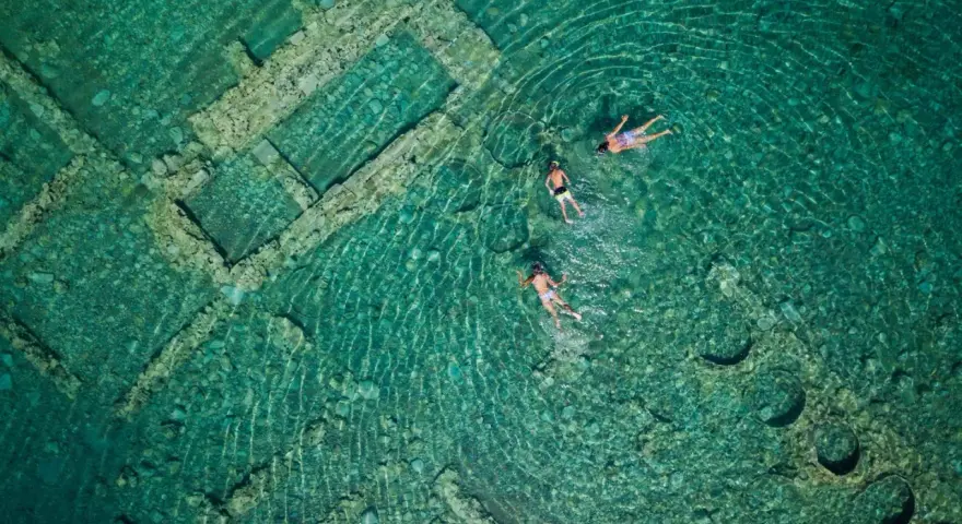 Swimmers explore the sunken city of Epidaurus, its ancient stone structures visible beneath the clear turquoise water.