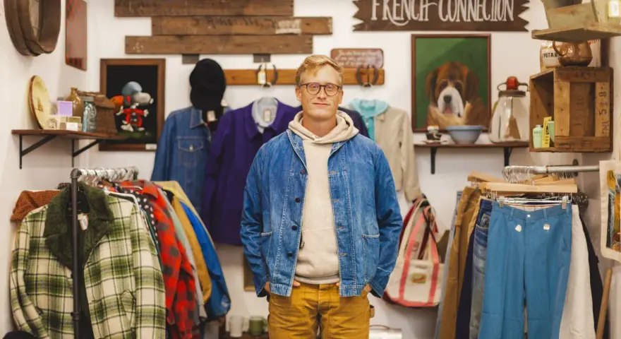 Mężczyzna w sklepie z odzieżą vintage, gdzie wisi szyld "FRENCH CONNECTION". W tle wiszą ubrania i dekoracje.