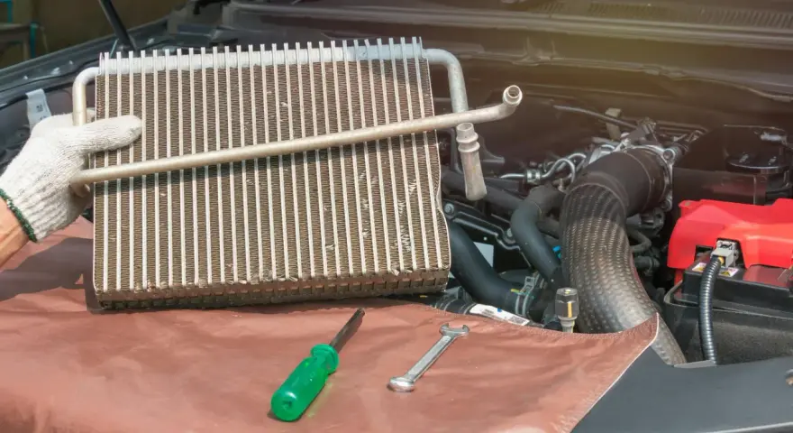 Gloved hand holds car AC evaporator. Tools and engine parts visible. Learn how to make AC colder in car by fixing this component.