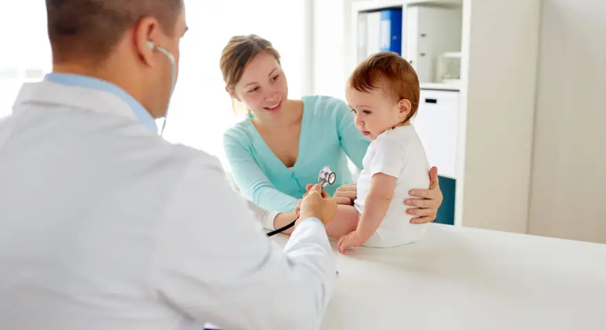 Pediatra bada niemowlę podczas pierwszej wizyty. Mama z uśmiechem obserwuje badanie.