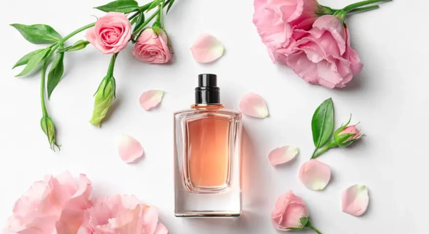 Elegancka butelka perfum z delikatnym, różowym płynem, otoczona pąkami i płatkami róż. Zapach jak słodka jeżyna.