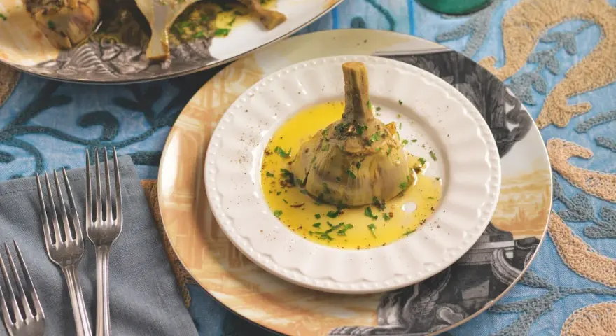 Un carciofo alla romana, tenero e profumato, adagiato su un piatto bianco con olio dorato e prezzemolo tritato.