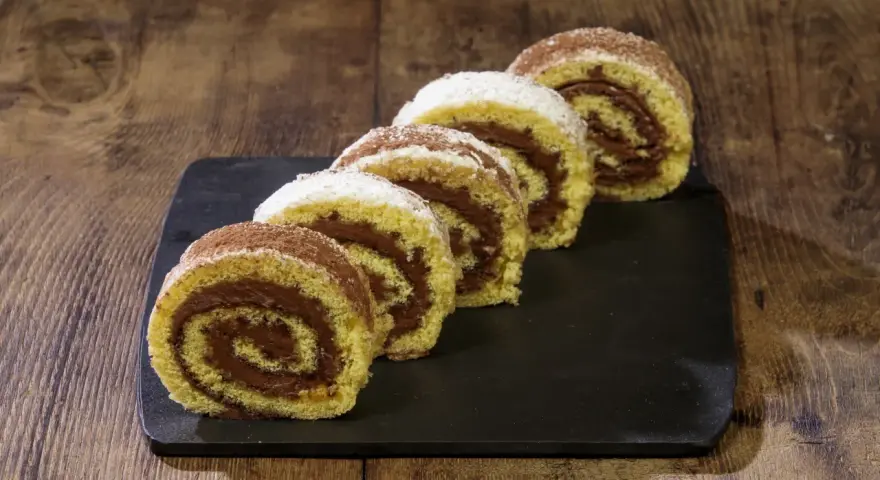 Fette di rotolo alla nutella cosparse di zucchero a velo e cacao, su una lavagna scura, pronte per essere gustate.
