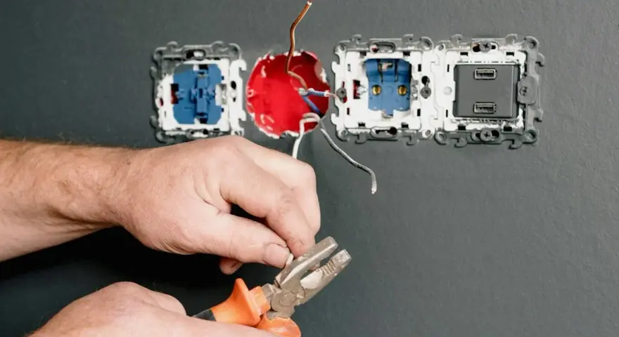 Dłonie trzymające kombinerki przy przewodach elektrycznych podłączanych do gniazdka.