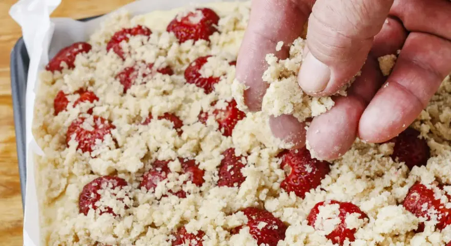 Ręka posypuje kruszonką ciasto z truskawkami. Zapowiada się pyszne ciasto z truskawkami i kruszonką!