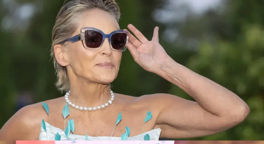Sharon Stone, con su elegante pelo corto con canas naturales, ajusta sus gafas de sol. Lleva un collar de perlas y un vestido adornado con plumas turquesas.