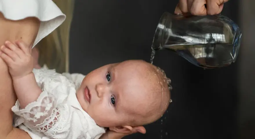 Chrzest święty. Woda spływa na główkę dziecka w białej szatce. Piękne życzenia na chrzest dla maluszka.