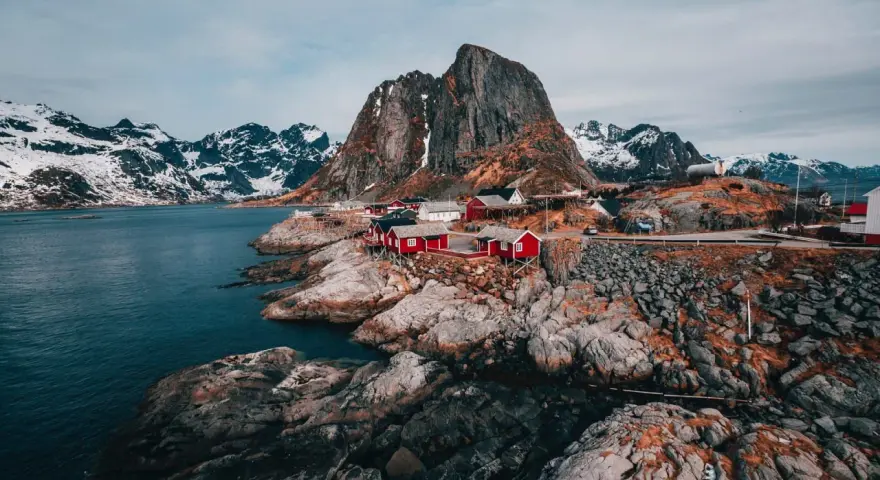 Röda fiskestugor vid en klippig kust med snöklädda berg i bakgrunden. En av de fina platserna i Norge.