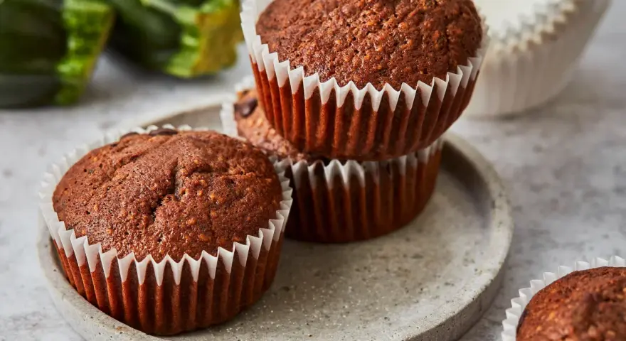 Saftige zucchini schoko muffins auf einem Steinplattenteller, im Hintergrund frische Zucchini.