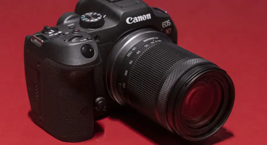 Appareil photo noir Canon EOS R7 avec objectif 18-150 mm, posé sur un fond rouge vif.