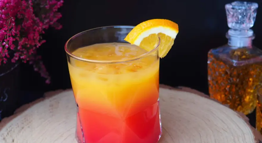 Kolorowy drink z koniakiem, ozdobiony plasterkiem pomarańczy, na tle karafki z bursztynowym alkoholem i kwitnących wrzosów.