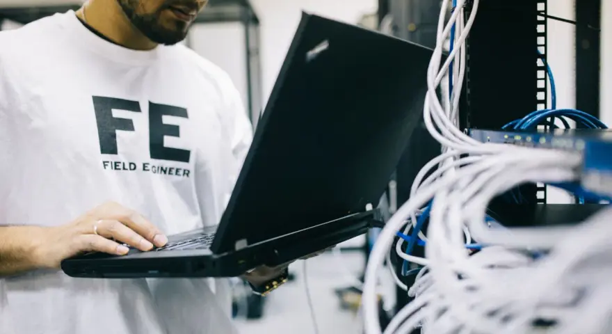 Inżynier w koszulce "FE FIELD ENGINEER" używa laptopa do sprawdzenia **trace route cmd** w serwerowni z plątaniną kabli.