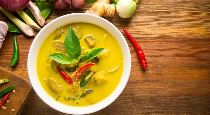 Aksamitne tajskie curry z tofu, ozdobione świeżą bazylią i papryczkami chili, otoczone składnikami jak galangal i trawa cytrynowa.