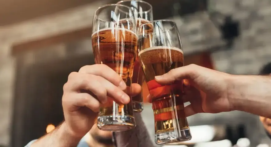 Trzy ręce unoszą kieliszki ze złotym piwem, tworząc toast. Każde piwo w szklance wygląda apetycznie.
