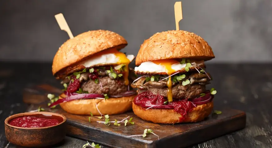 Zwei saftige Burger mit Spiegelei, Pilzen und roten Zwiebeln, serviert auf einem Holzbrett. Ein Muss für jeden Burger Laden in Berlin.