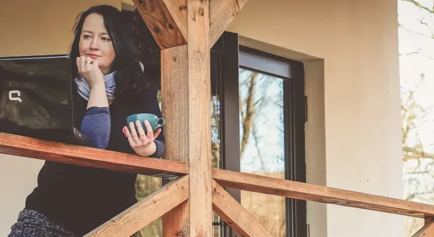 Kobieta z kubkiem kawy pracuje na laptopie na werandzie. Internet na działce umożliwia jej pracę w otoczeniu natury.