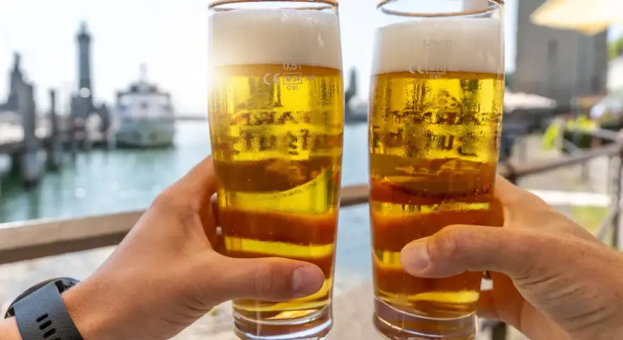Zwei Hände halten Biergläser mit schaumigem Bier. Im Hintergrund ein Hafen. Ist Bier gesund? Ein Genuss am Wasser.