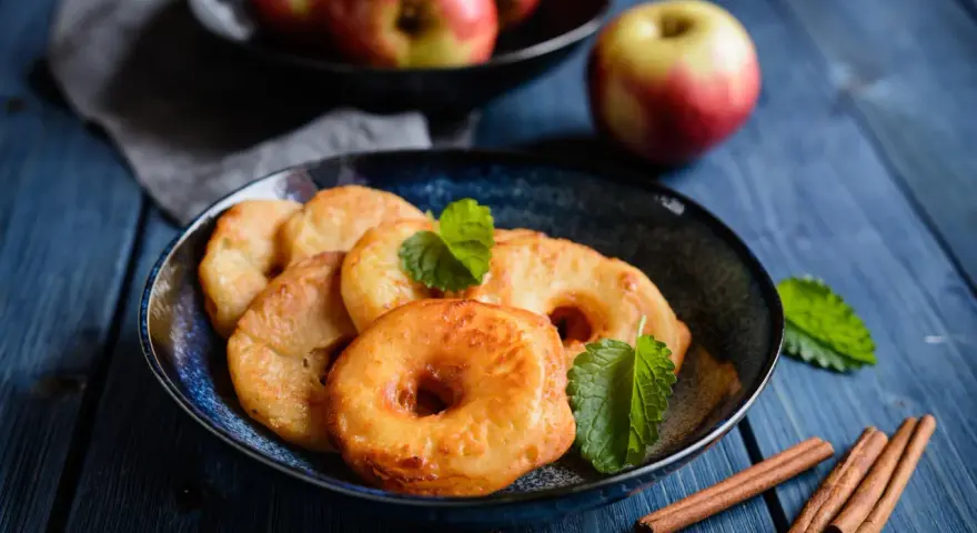 Délicieux beignets aux pommes dorés, garnis de menthe fraîche et accompagnés de bâtons de cannelle sur une table en bois bleu.