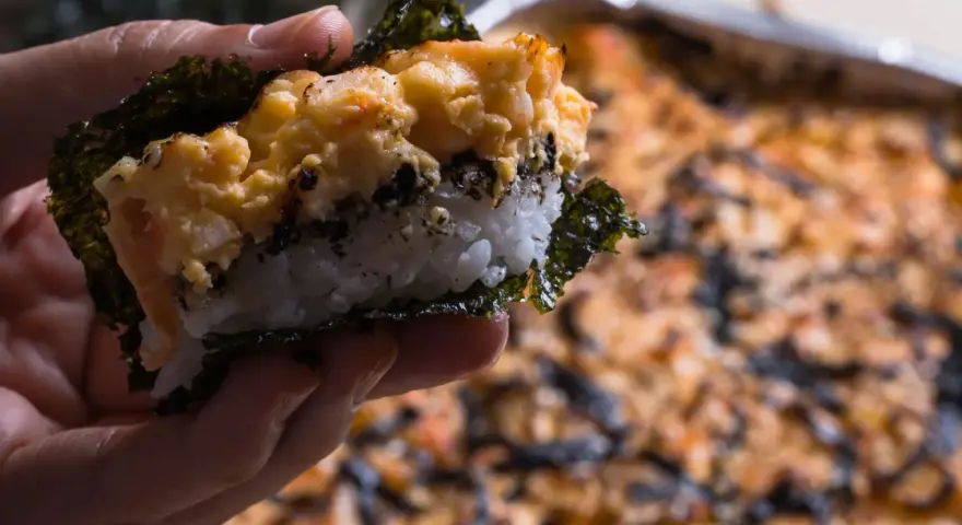 Dłoń trzyma kawałek sushi bake z ryżem, nori i zapiekanym farszem.