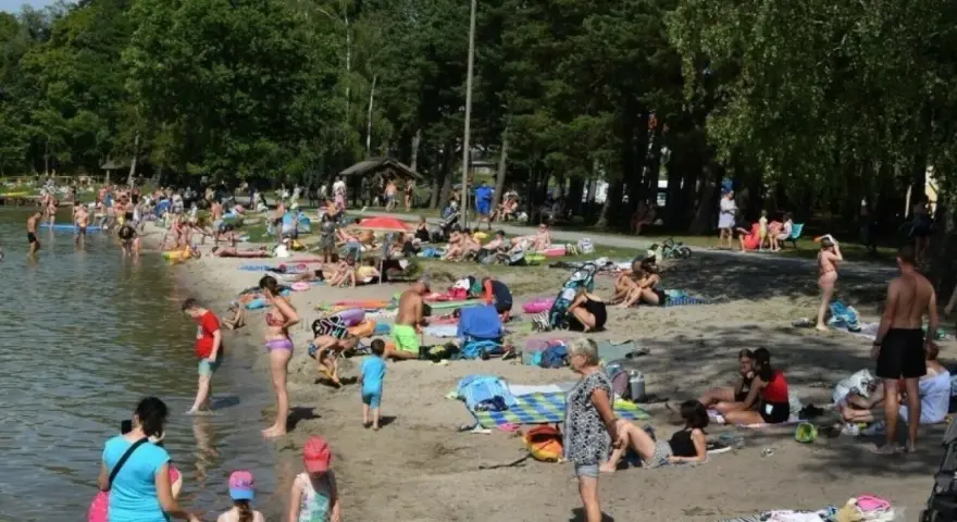 Tłoczna plaża nad jeziorem na Pomorzu. Ludzie opalają się, kąpią i odpoczywają w cieniu drzew.