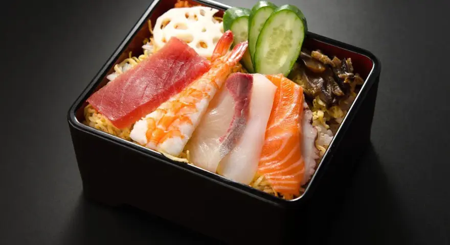 Chirashi zushi: kolorowe plastry ryb (tuńczyk, łosoś, biała ryba), krewetka, plasterki ogórka i lotosu na ryżu.