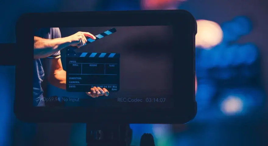Ręka zamyka klaps filmowy. Na ekranie widać napis "REC:Codec 03:14:07". To plan średni, zapowiadający rozpoczęcie nagrania.