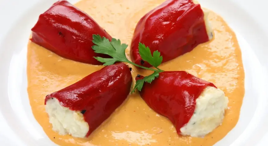 Cuatro pimientos rojos rellenos de bacalao, servidos sobre una cremosa salsa naranja y decorados con perejil.