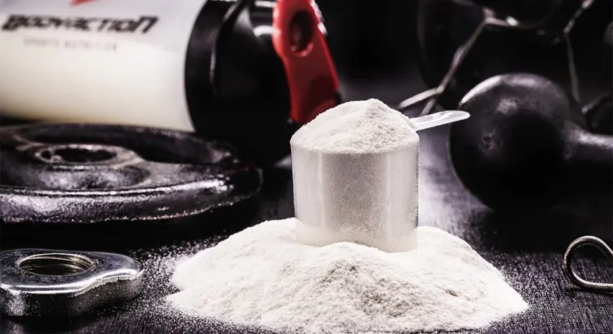 Polvo blanco en una cuchara medidora, junto a pesas y un bote de suplemento. Descubre para qué sirve la creatina y potencia tus entrenamientos.