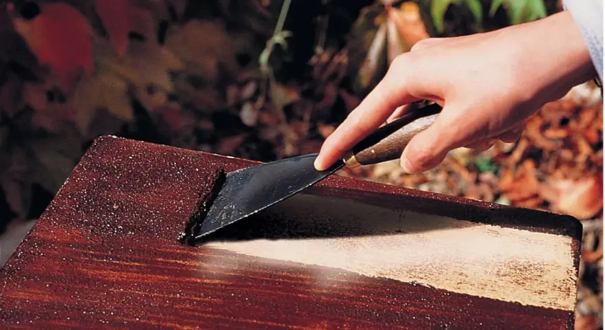 Main d'une personne utilisant un grattoir pour enlever de la peinture d'un meuble en bois, comme avec un décapant peinture bois fait maison.