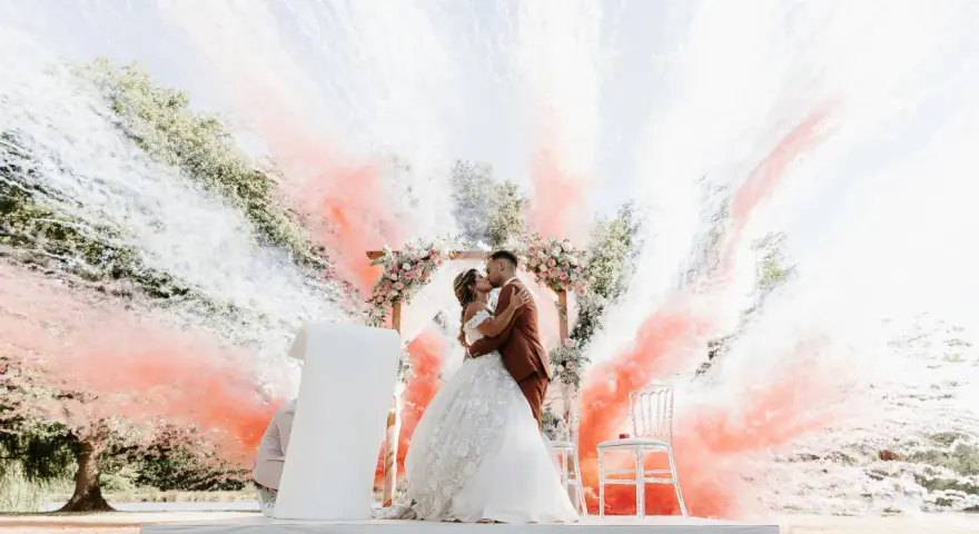 Un couple s'embrasse sous une arche fleurie, entouré de fumée rose et blanche. Ces photos mariage capturent un moment magique.