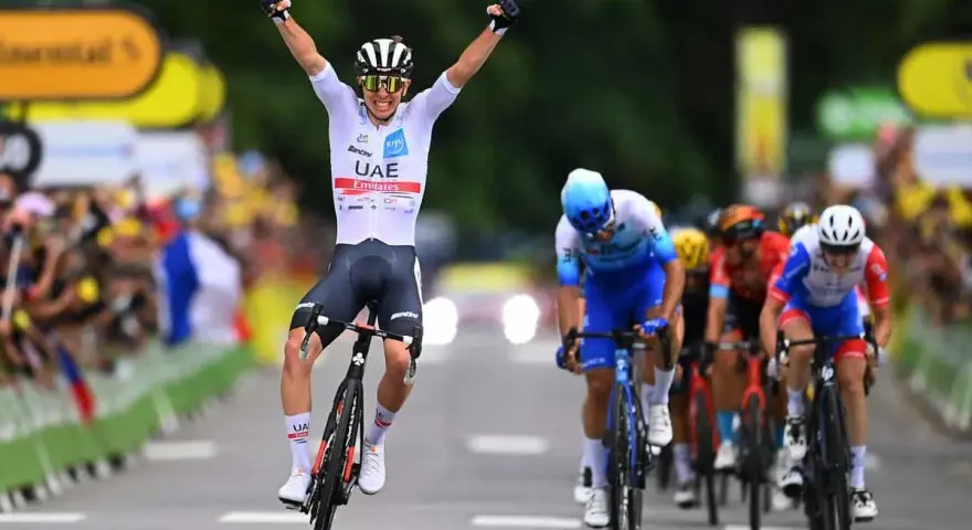 A cyclist in a white UAE Emirates jersey celebrates victory at the Tour de France, arms raised in triumph as he crosses the finish line.