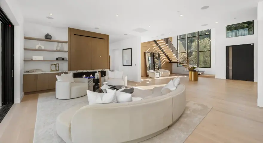 Soggiorno luminoso con camino moderno, divano curvo e poltrone bianche. Scale a vista e grandi finestre definiscono l'arredamento minimal chic.