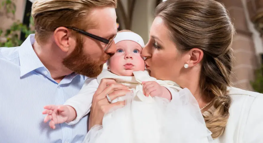 Eltern küssen ihr Baby bei der Taufe. Die Frage, wie viele Paten darf man haben, beschäftigt viele.