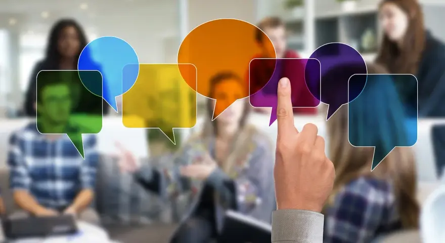 Ludzie w tle, kolorowe dymki dialogowe symbolizujące komunikację. Dłoń wskazująca na jeden z dymków, sugerująca aktywny udział w dyskusji. To obrazuje pracę zespołu interdyscyplinarnego.