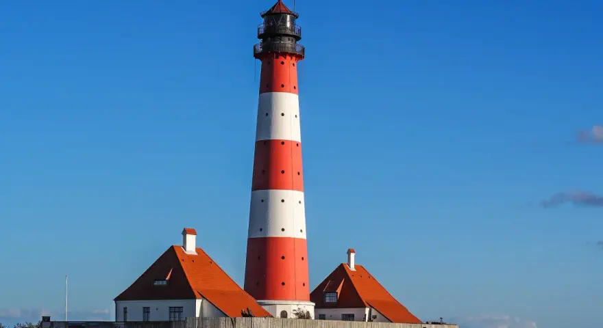 Wysoka, czerwono-biała latarnia morska w Jastarni strzeże wybrzeża. Obok stoją dwa domki z pomarańczowymi dachami.